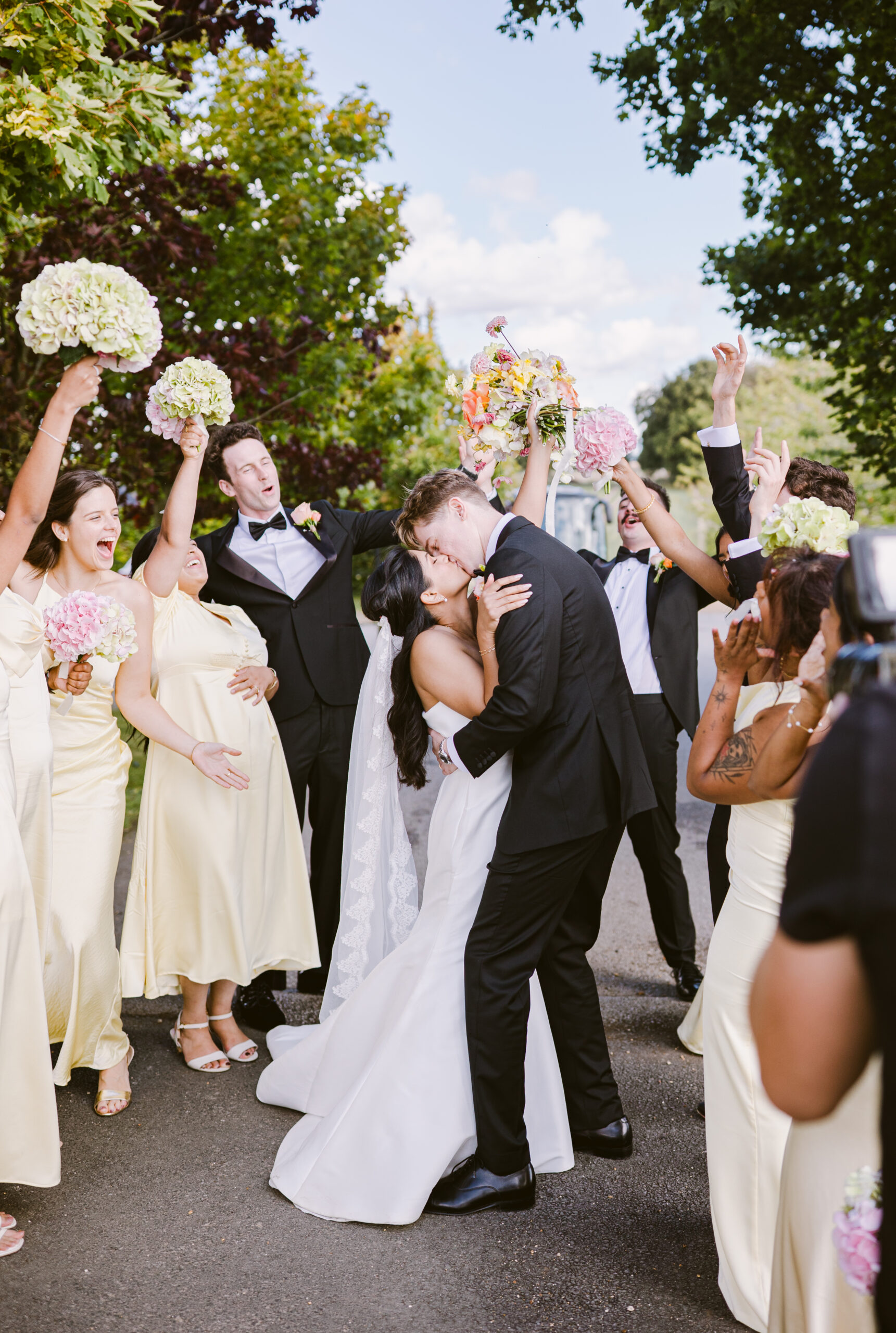 bride and groom kissing whilst their bridal party are waving their hydrangea flowers around and cheering them on