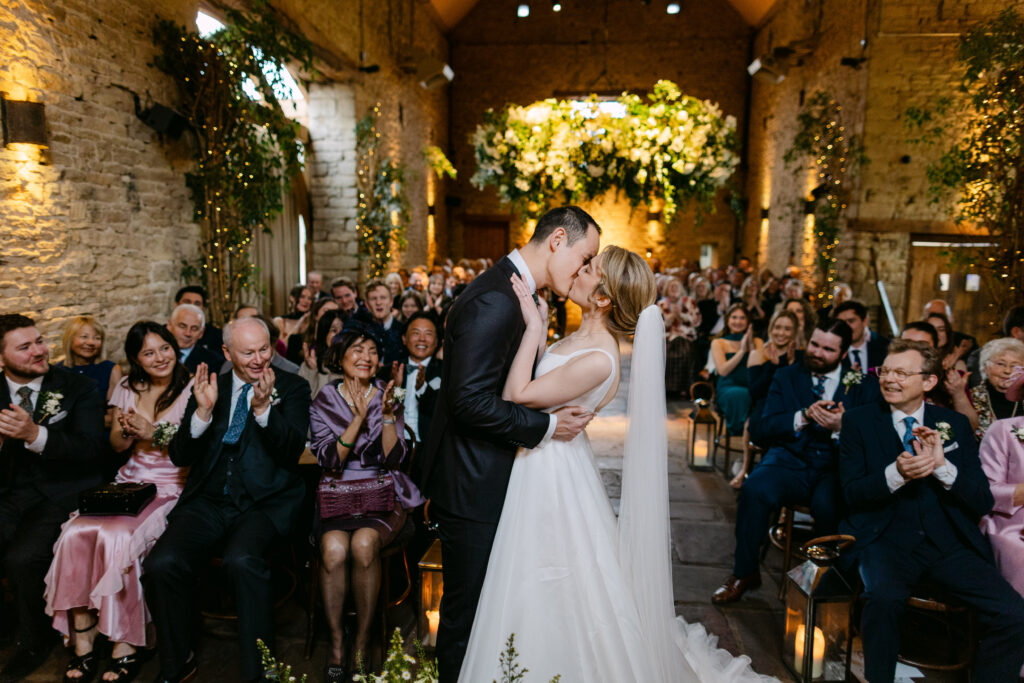 bride and groom having first kiss at cosy winter wedding at cripps barn. guests clapping