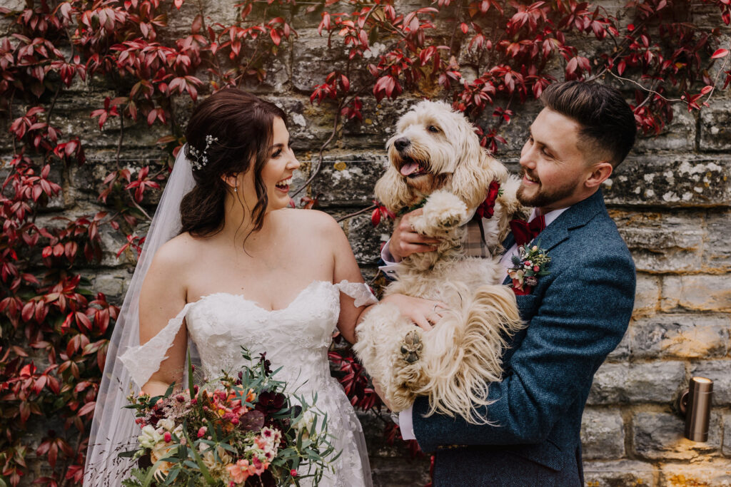 Bride and groom holding their dog during an autumn wedding at Elmore Court, framed by red ivy on a stone wall.
