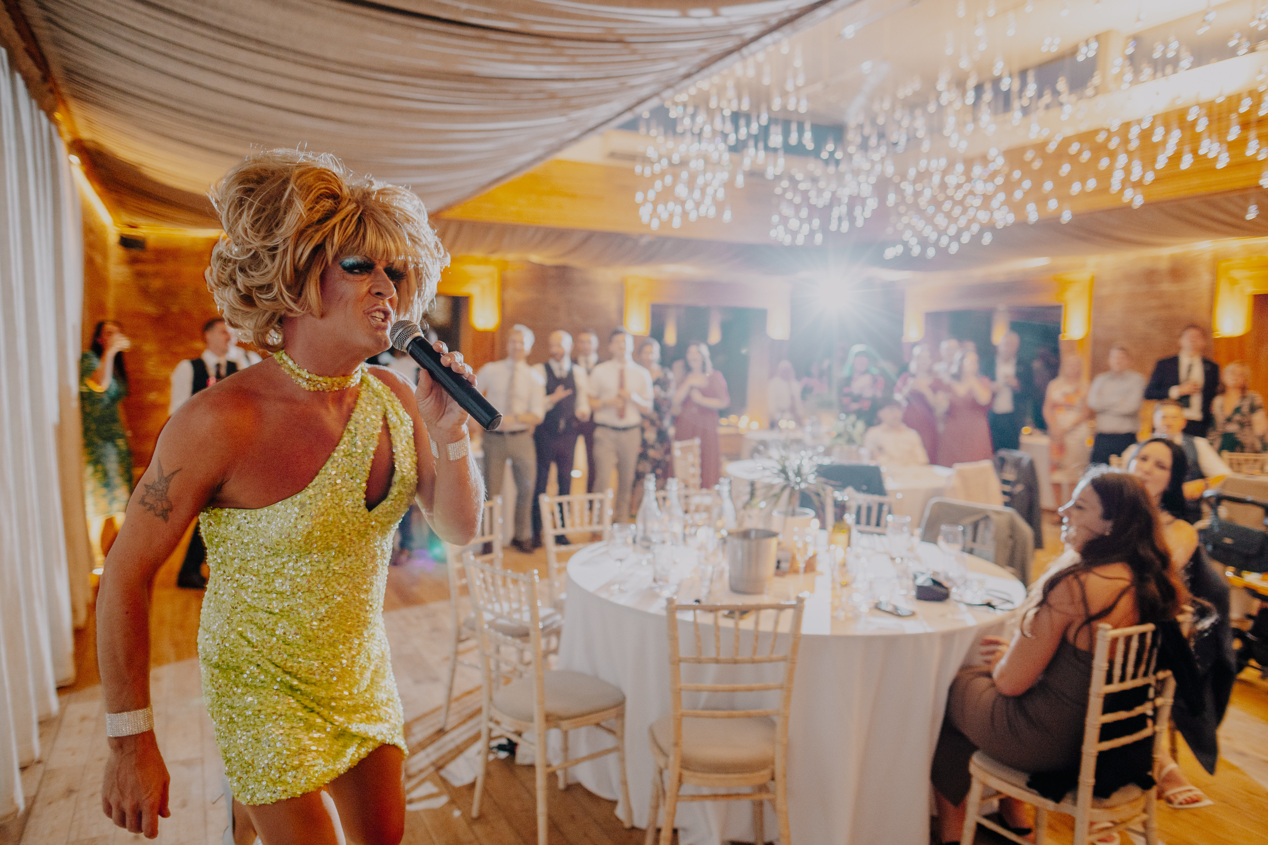 drag queen singing to the party in the gillyflower with a big wig on and yellow sequin dress