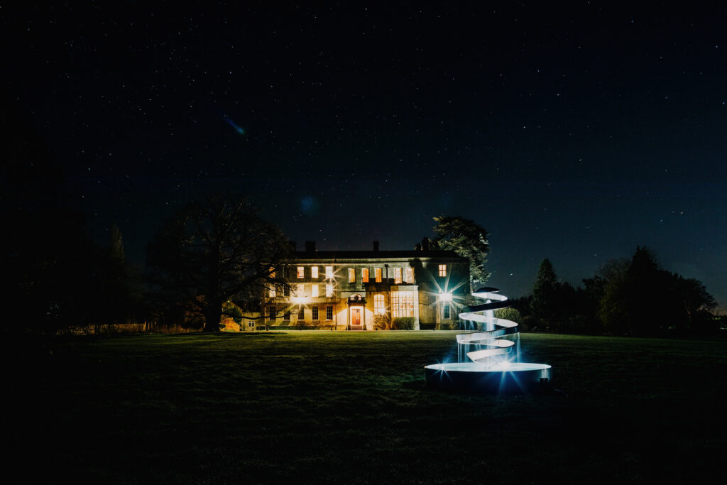Elmore Court illuminated at night, with the house glowing under a starry sky and sculpture in the foreground.