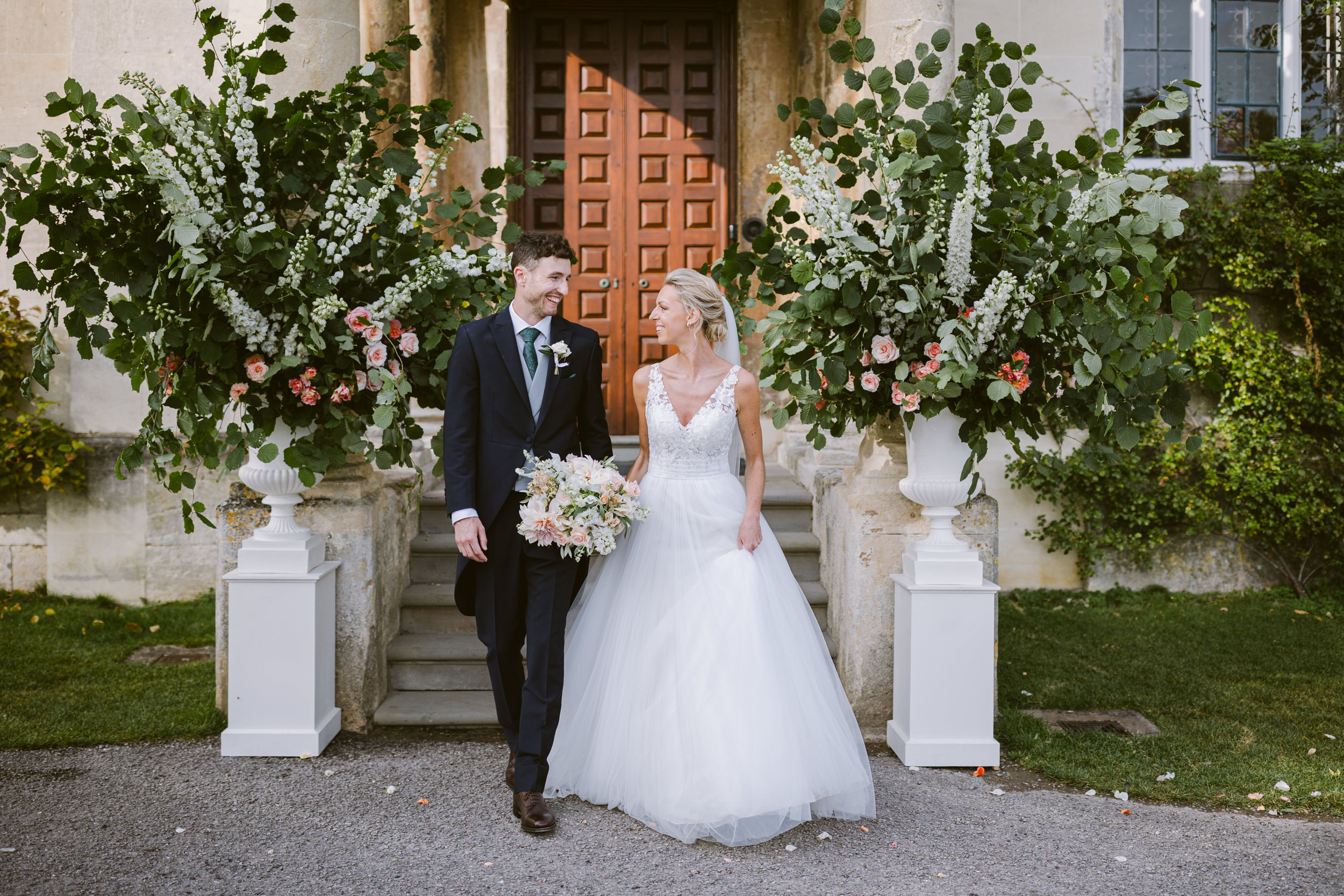Bride and groom walking together outside Elmore Court in early autumn, surrounded by greenery and soft seasonal light.