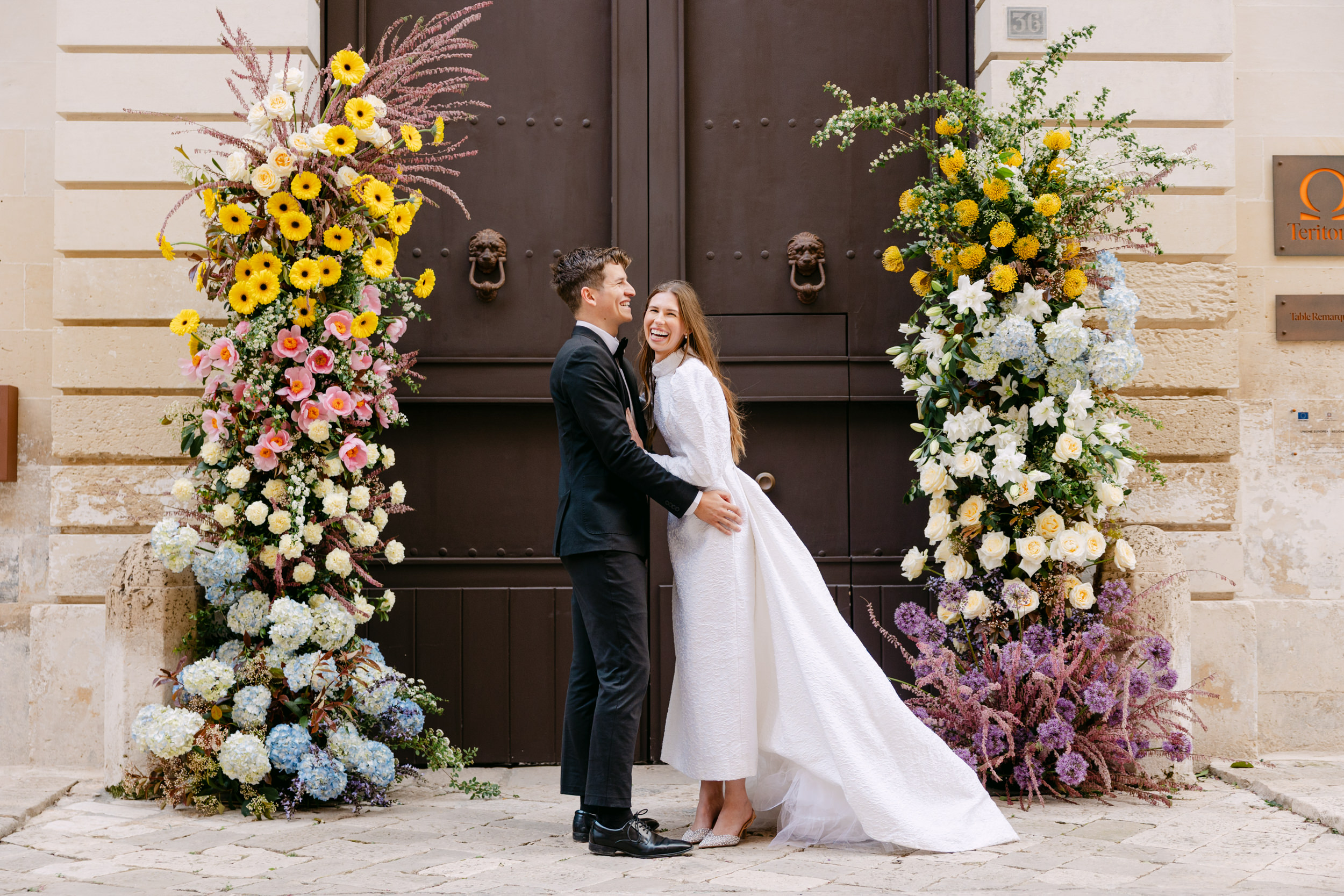 bride and groom laughing together standing in front of old Italian doors surrounded by wedding flowers at a puglian wedding