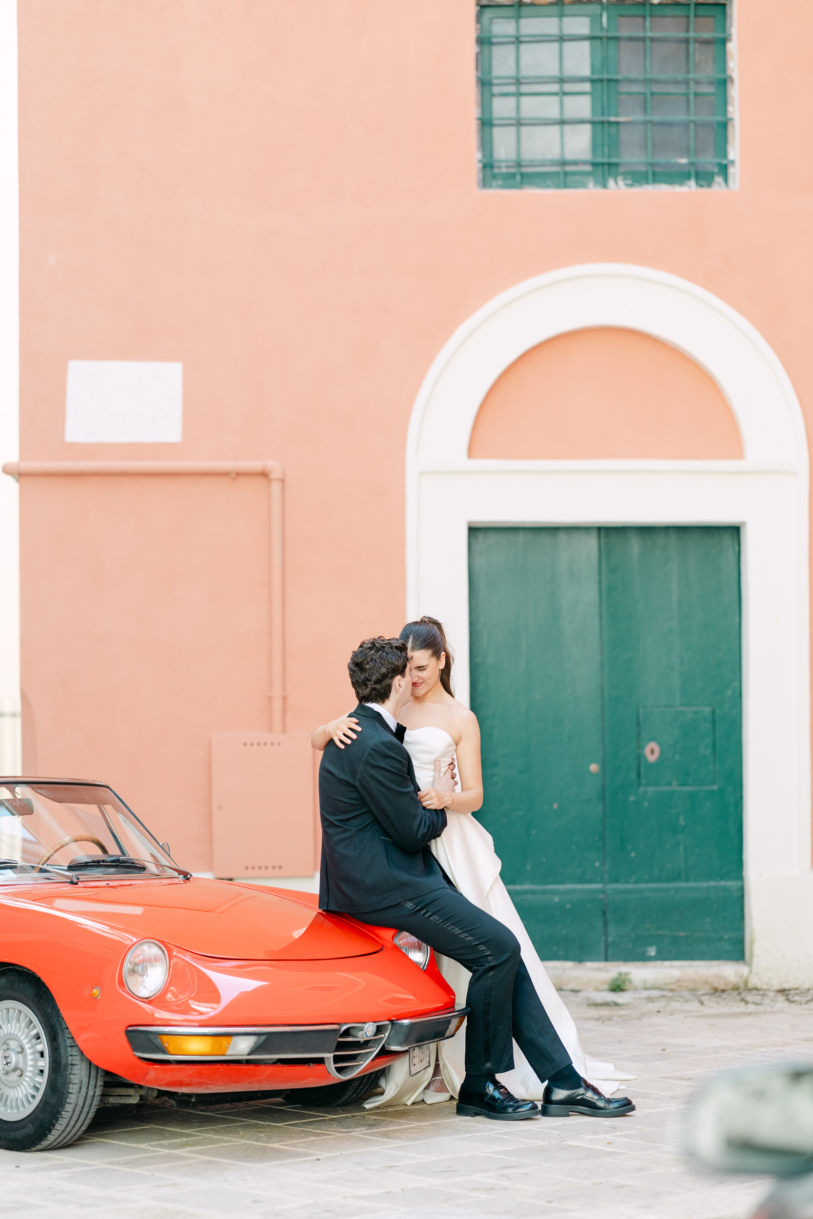 groom sitting on a vintage alfa Romeo with is back to the camera and cuddling his bride